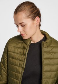Doudoune vert olive avec col montant, dotée d'une fermeture éclair et de coutures horizontales. Portée par-dessus un haut noir. Texture lisse.