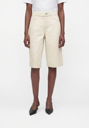 Person iført knælange beige shorts, hvid kortærmet skjorte og sorte spidse hæle, står mod en hvid baggrund.