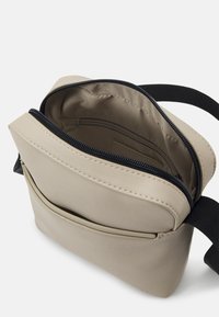 Beige formlæder taske med en lynlåslukket hovedkompartment og en udvendig lomme, der har en sort rem og glat tekstur.