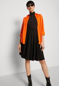 Camicia arancione con bottoni, dolcevita nera e gonna plissettata nera. Il modello indossa occhiali da sole neri e stivaletti alla caviglia. Tessuto liscio con una vestibilità su misura.
