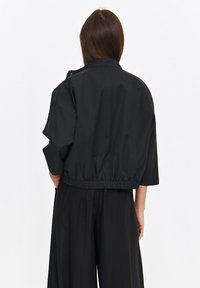 Przycięta czarna kurtka z wysokim kołnierzem, luźnym krojem, elastycznym paskiem w talii i krótkimi rękawami, wykonana z gładkiego, lekkiego materiału.