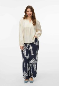 Blusa color crema con una finitura testurizzata, dettagli a bottoni, abbinata a pantaloni blu navy con audaci motivi geometrici bianchi. Scarpe blu.