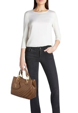 Borsa marrone trapuntata con dettagli dorati, etichetta a forma di cuore e manici superiori. Indossata con una camicia bianca e jeans neri, presenta un design elegante.