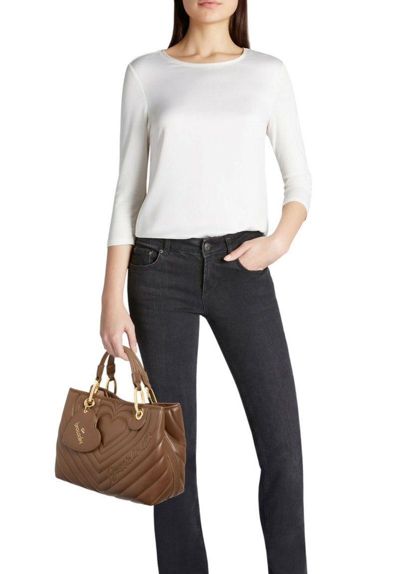 Borsa marrone trapuntata con dettagli dorati, etichetta a forma di cuore e manici superiori. Indossata con una camicia bianca e jeans neri, presenta un design elegante.