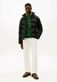 Zwarte donzen jas met gewatteerd ontwerp en ritssluiting, gedragen over een groene trui, gecombineerd met witte broek en bruine schoenen. Effen achtergrond.