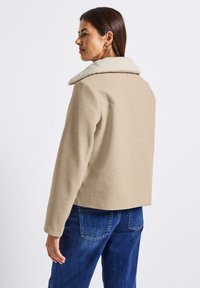 Krémová zkrácená bunda s jemným krémovým, fleecem podšitým límcem. Má dlouhé rukávy, jednoduchý design a je kombinována s modrými džínami.