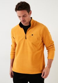 Žuti flis pulover s polu-zip ovratnikom, crnim akcentima i logotipom na prsima. Ima dugačke rukave i mekan, teksturiran završetak.