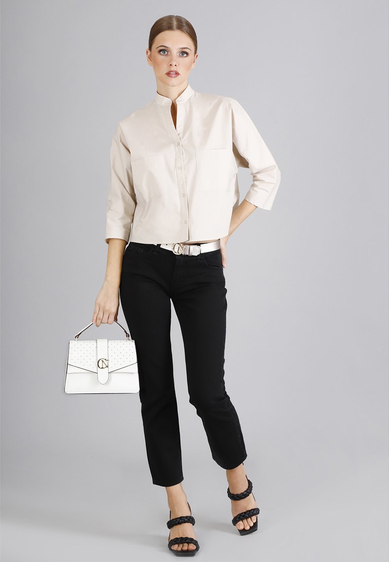 Camicia beige corta con colletto stand, jeans neri, tacchi intrecciati neri e una borsa bianca con accento dorato e dettaglio a pois.