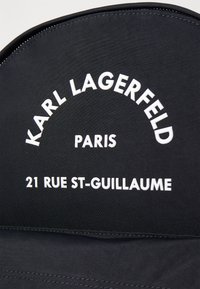 Textile noir avec du texte blanc en relief : "KARL LAGERFELD," "PARIS," et "21 RUE ST-GUILLAUME," avec une texture lisse et une forme structurée.