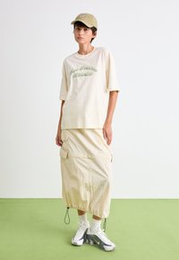 Beige oversized t-skjorte med grønn "New Balance Athletics" brodering, parret med et matchende beige stoffskjørt, sokker i kremfarge og grå sneakers.