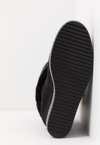 Botte d'hiver noire avec semelle nervurée face à la caméra, affichant la taille 37 sur le talon, positionnée contre un mur d'angle blanc.