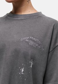 Szary bawełnowy sweter z logo i tekstem wyhaftowanym na biało, z plamami na przodzie. Okrągły dekolt i luźny krój.