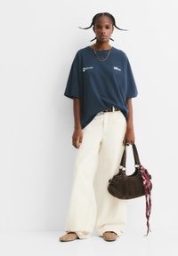 Marineblå oversized t-shirt, cremefarvede vide jeans, brun håndtaske med et tørklæde og beige slip-on sko med perforeringer. Står på en hvid baggrund.