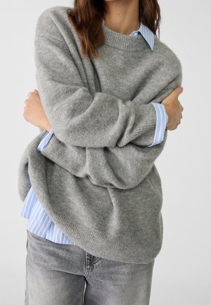 Persoon draagt een losse grijze trui over een blauw-wit gestreept shirt, met gekruiste armen, gecombineerd met vervaagde grijze spijkerbroek.