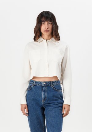 Cropped white button-up shirt with long sleeves, two front pockets, soft texture, worn with high-waisted blue jeans.