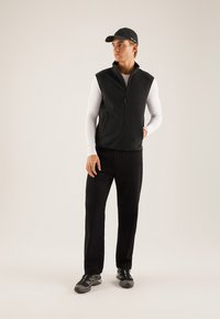 Black fleece vest with a zip closure, paired with a white long-sleeve shirt, black pants, and black sports shoes. Cap completes the look.