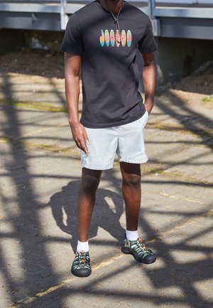 Person iført en sort Jordan t-shirt, lysegrå shorts, hvide sokker og grønne udendørs sandaler, stående på en skyggefuld fortov.