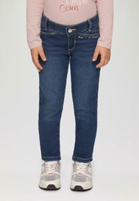 Niebieskie dżinsy w slim fit, z przednimi kieszeniami, metalowym zapięciem na guzik i dekoracyjnym przeszyciem na kieszeni. Noszone z szarymi sneakersami.