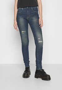 Vaqueros ajustados de mezclilla de color azul, con áreas desgastadas, una cintura de altura media y botas negras de cordones con una suela gruesa que complementan el conjunto.