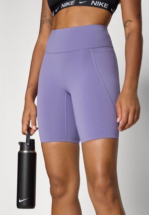 Frau trägt lila Biker-Shorts mit hoher Taille und schwarzen Nike Sport-BH, hält eine schwarze Nike Wasserflasche an ihrer Seite.