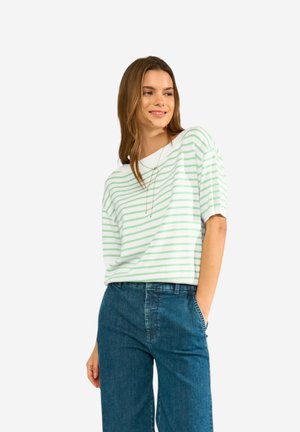 Woman smiling, wearing white and light green striped short-sleeve top and blue high-waisted jeans, standing with one hand in pocket.