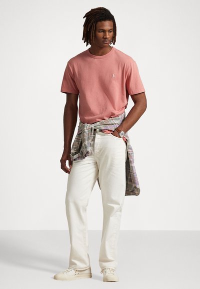 Roze katoenen T-shirt, relaxte fit, met klein logo. Crème-kleurige spijkerbroek en een geruit shirt om de taille geknoopt. Witte sneakers.