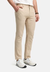 Chinos de algodón beige con un ajuste slim, bolsillos frontales y detalles de costura visibles, combinados con zapatillas blancas con acentos grises.