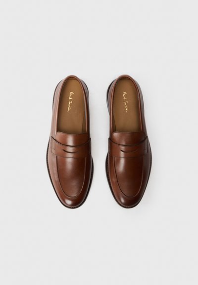 Mocasines de cuero marrón con un acabado suave, costuras sutiles y una banda decorativa en la parte superior, sobre un fondo liso.