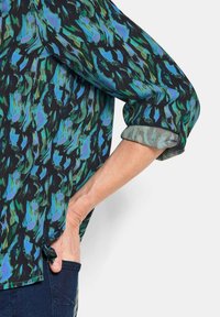 Luftige Bluse mit schwarzem Hintergrund und einem grünen und blauen abstrakten Muster; die ärmellosen Manschetten zeigen einen leichteren, kontrastierenden Stoff darunter.