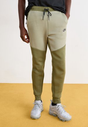 Le pantalon de survêtement vert Nike présente un design coloré avec des nuances de vert plus claires et plus foncées, une taille élastique et des poches latérales, associé à des chaussures argentées.