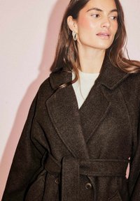 Brūns vilnas mētelis ar šallē līdzīgu apkakli, jostotu vidukli un lieliem pogām. Mīksta tekstūra un brīvs piegriezums. Modelis valkā gaišu krekla zempusi.