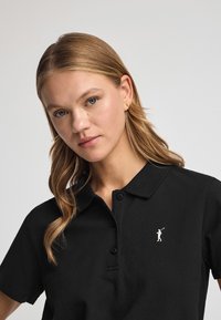 Polo noir avec un col boutonné, comportant un petit logo blanc sur la poitrine. Tissu doux et texturé avec une coupe ajustée.
