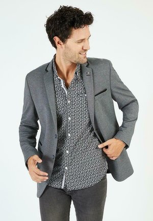 Blazer gris avec une finition texturée, mettant en valeur une accent de poche noire et des boutons, superposé à une chemise à motifs noir et blanc.
