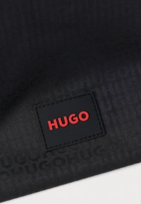 Čierna texturovaná tkanina s jemným vzorom slova „HUGO“. Výrazná obdĺžniková čierna etiketa s červeným nápisom „HUGO“.