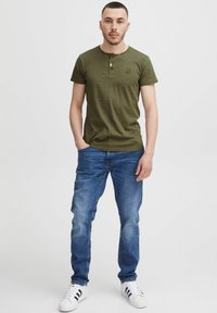 Olivgrünes kurzärmeliges T-Shirt mit Henley-Ausschnitt und kleinem Logo, kombiniert mit blauen Jeans und weißen Sneakers mit schwarzen Streifen.