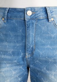 Ljust blå denimjeans med ett urblekt mönster av små abstrakta mönster. Har en knappstängning och metallaccenter i midjan.