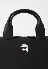 Czarna torebka z gładkiego materiału, zaokrąglonymi uchwytami, zamkiem błyskawicznym oraz wyróżniającą się czarno-białą naszywką z wizerunkiem twarzy w okularach przeciwsłonecznych.