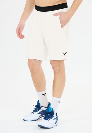 Mand iført hvide sportsshorts med sort taljebånd, hvide sokker og hvide og blå sportssko, stående mod hvid baggrund.