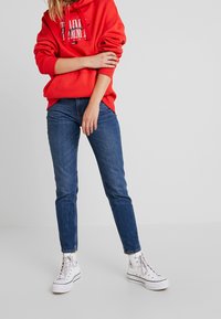 Person trägt einen roten Kapuzenpullover, blaue Jeans und weiße High-Top-Sneaker und steht vor einem schlichten hellgrauen Hintergrund.