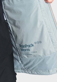 La giacca impermeabile azzurra presenta all'interno l'etichetta "RainPack Warm", icone che indicano il controllo della temperatura e una texture liscia. Chiusura con zip visibile.