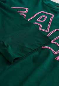 Grünes Baumwoll-T-Shirt mit pinken gestickten Buchstaben, mit glatter Textur und umgeschlagenem Ärmel, der klare Stichdetails zeigt.
