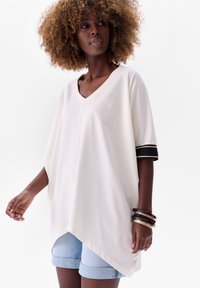 White oversized V-neck top with short sleeves featuring black and gold accents, paired with light denim shorts and multiple bangles.