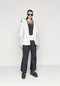 Síruha, amely egy fehér és világoskék kapucnis kabátból, fekete nadrágból és fekete síbakancsból áll. A megjelenést napszemüveg teszi teljessé.