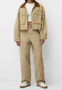 Chaqueta recortada beige con cuello de pana marrón, dos bolsillos frontales; combinada con pantalones de corte holgado a juego, con detalles de cremallera y pernera ancha.