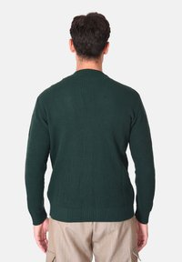 Maglione verde a coste con maniche lunghe, scollo rotondo, maglia strutturata e orlo aderente. Indossato sopra pantaloni beige con tasche laterali.