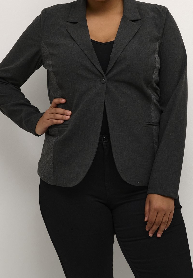 Femme portant un blazer gris foncé cintré sur un top noir et un pantalon noir, posant avec une main sur la hanche devant un fond uni.