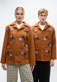 Καφέ σακάκια από μείγμα μαλλιού με κεντήματα με λουλούδια σε ροζ και πράσινο, με δύο μπροστινές τσέπες και γιακά, φορεμένα από δύο μοντέλα.