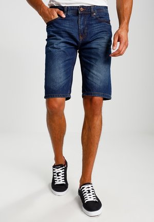 Mørkeblå denimshorts med klassisk pasform, optrækkede kanter og fem-lommer design. Båret med sorte sneakers og en hvid skjorte.