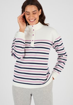 Femme souriante portant un pull blanc avec des rayures horizontales roses et bleu marine et un pantalon gris, touchant sa tempe d'une main.
