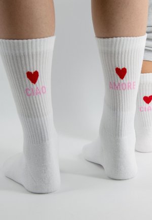 Chaussettes blanches à côtes portées aux pieds, chaque chaussette présente un cœur rouge et un texte rose indiquant "CIAO" et "AMORE" à l'arrière du mollet.